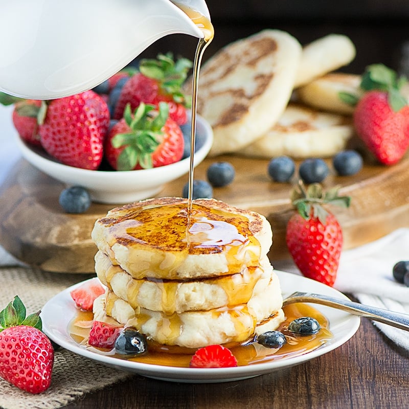 side view of 3 ricotta pancakes stacked on a white plate drizzled with syrup with berries in the background