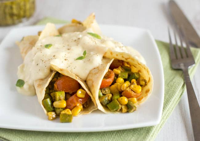 Veggie stuffed savoury pancakes with feta cheese sauce by amuse your bouche rolled up on a white plate with a fork and green napkin on the side.