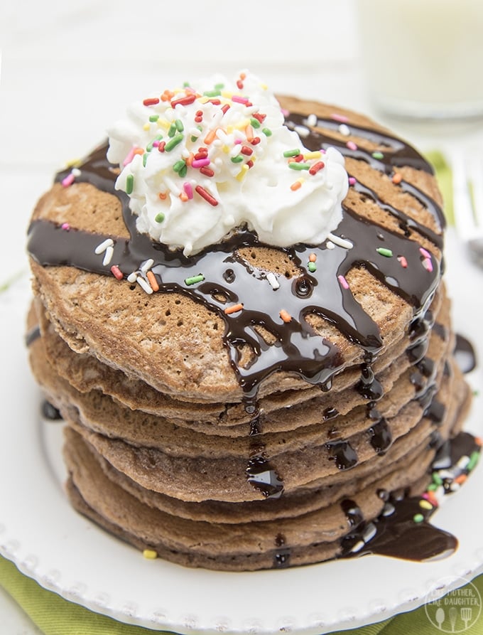 brownie batter pancakes by like mother like daughter stacked on a white plate with chocolate sauce drizzled on top with sprinkles and cream.
