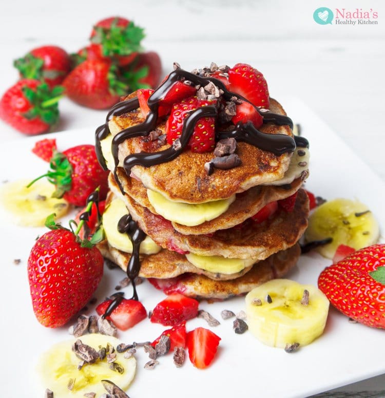 strawberry chocolate chip vegan pancakes by nadia's healthy kitchen stacked on a white plate surrounded by berries, bananas and chocolate sauce.