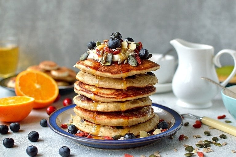 a stack of superfood pancakes topped with berries, yogurt and seeds on a blue plate with fruit and a milk jug in the background.