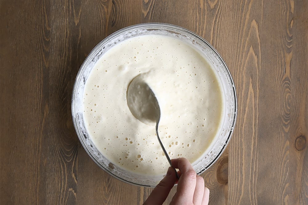 The vanilla sponge batter with a spoon held by a hand in a clear bowl.