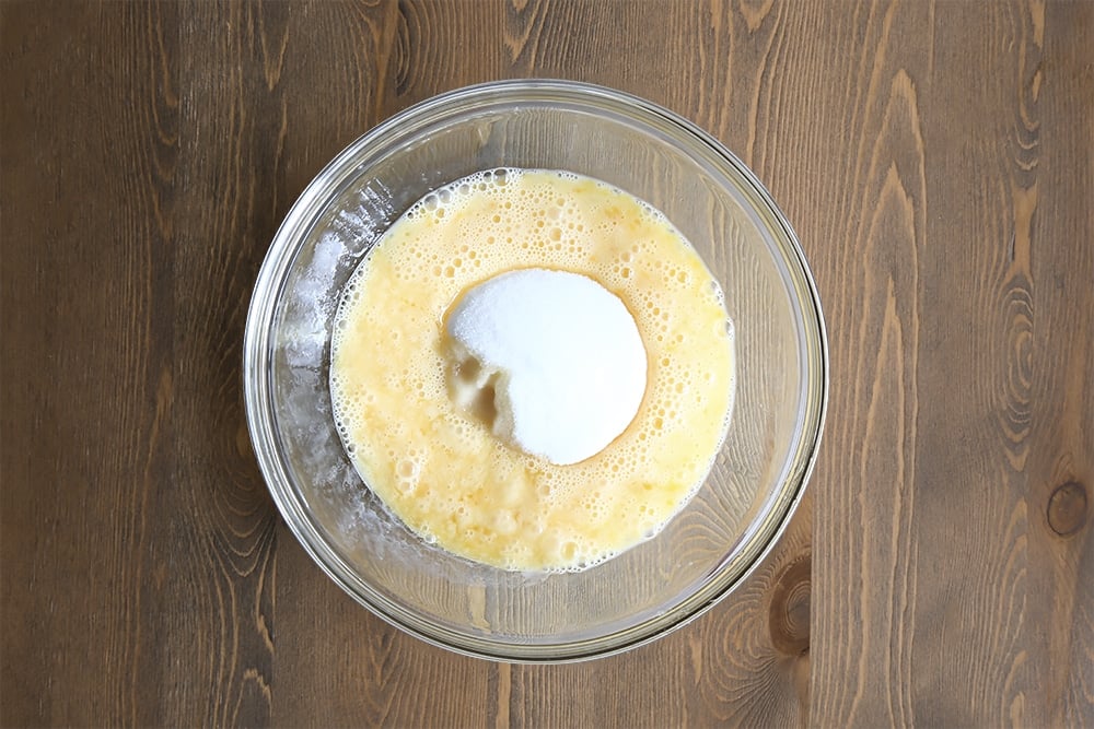 Sugar and eggs are mixed together in a clear bowl on a wooden background