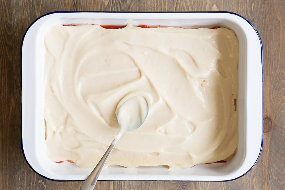 white metal dish with a strawberry mixture topped with a thin layer of vanilla sponge and white egg mixture with a spoon on top