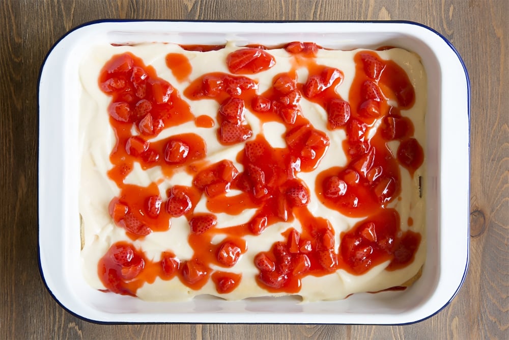 white metal dish with a strawberry mixture topped with a thin layer of vanilla sponge and white egg mixture and topped with a strawberry jelly layer.