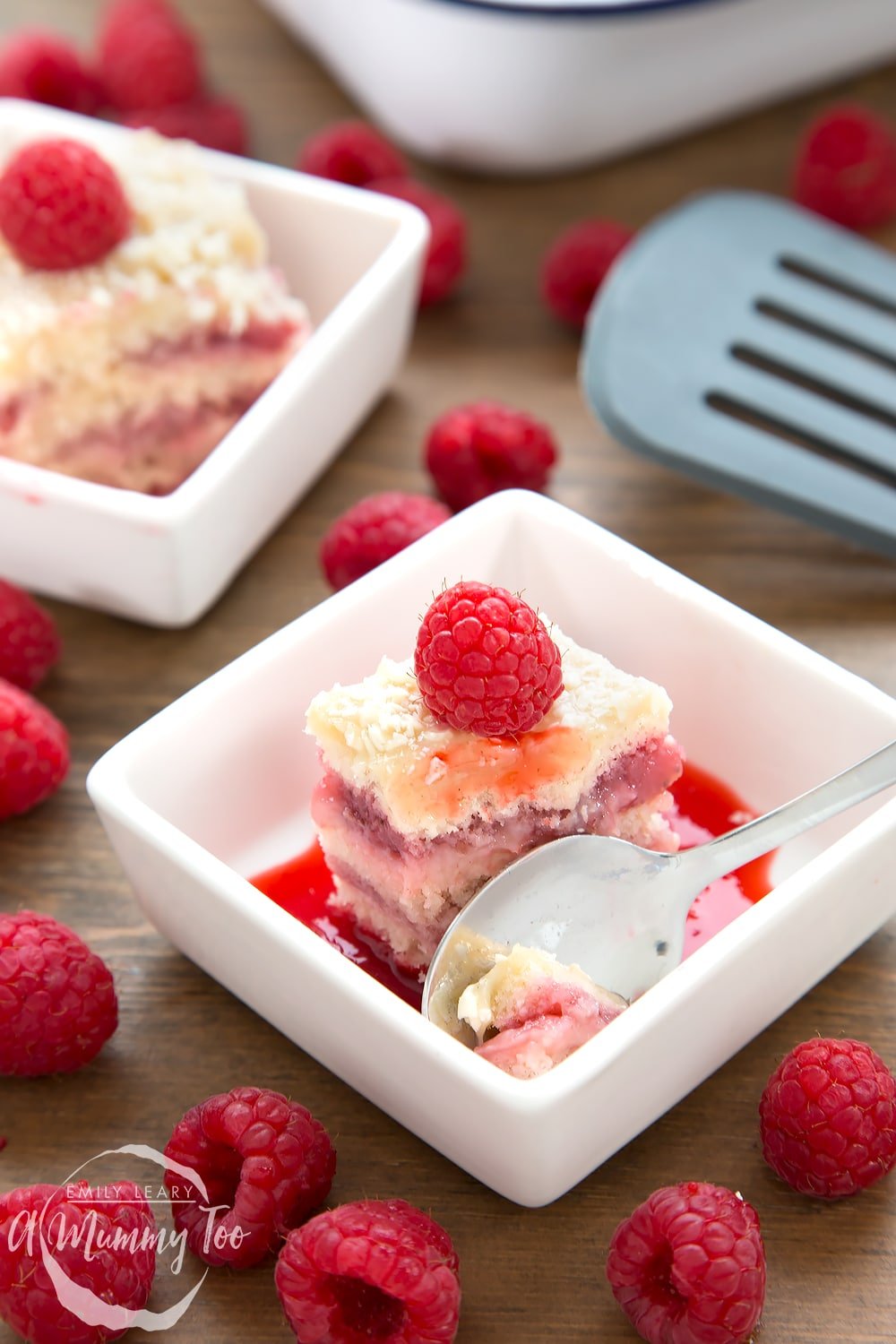 A piece of Strawberry and custard layer cake in a square white bowl, spoon on the side topped with a raspberry.