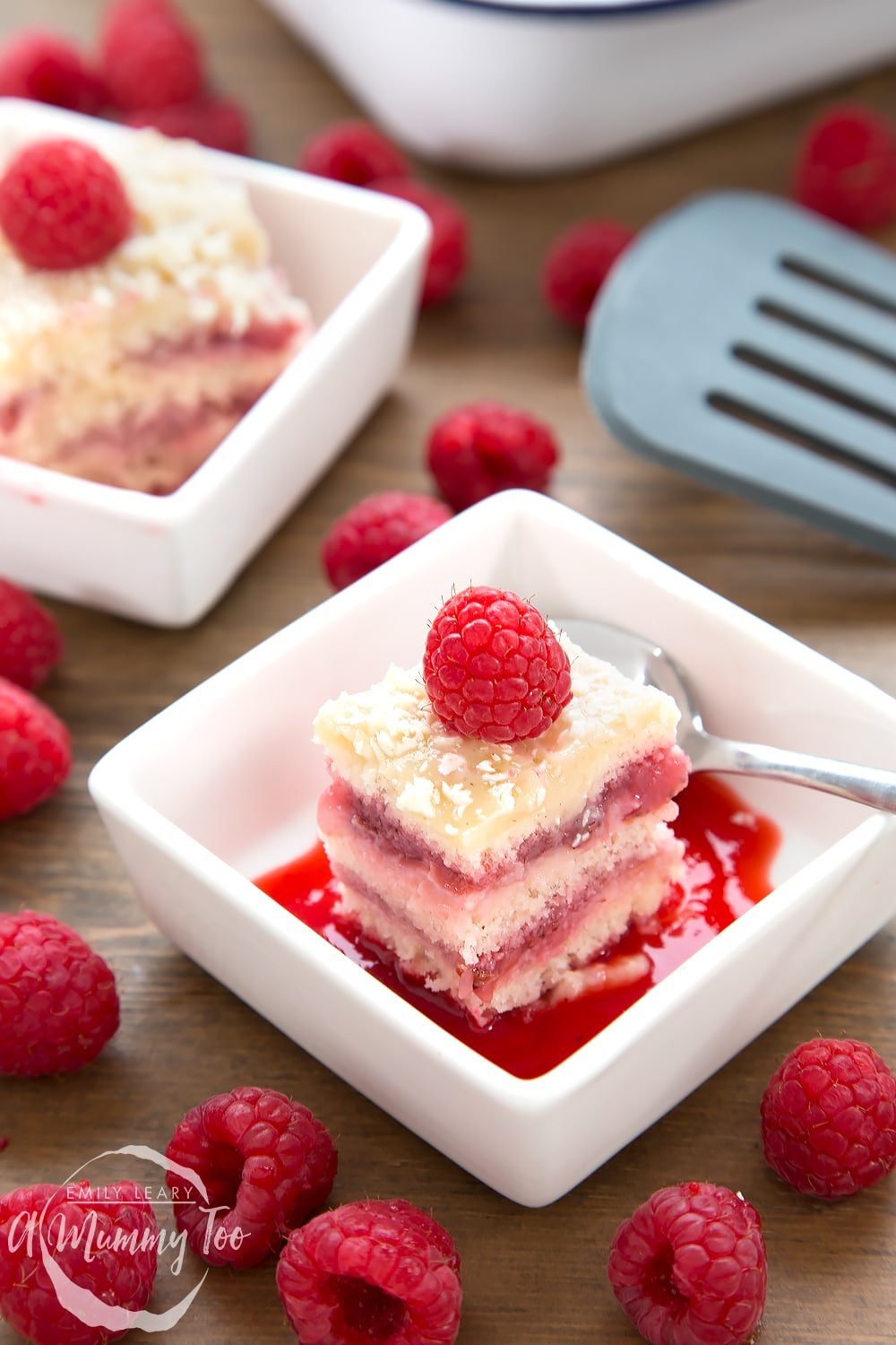 Strawberry layer cake square topped with a raspberry in a square white bowl and spoon on the side.