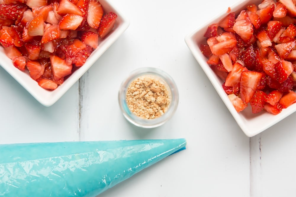 over head view of a small jar with crushed biscuit crumbs in the base with two bowls of chopped strawberries at either side and a blue icing bag.