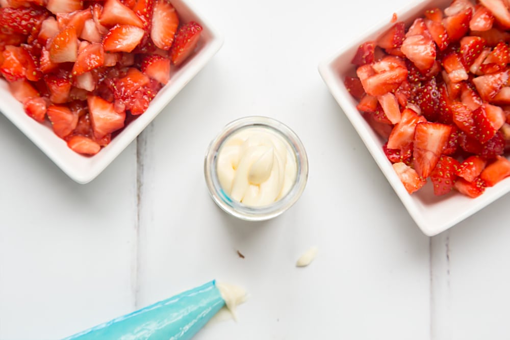 over head view of a small jar with crushed biscuit crumbs in the base and icing on top with two bowls of chopped strawberries at either side and a blue icing bag.