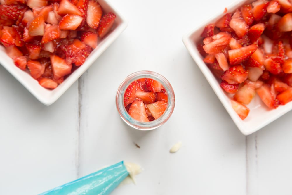 over head view of a small jar with shopped strawberries with two bowls of chopped strawberries at either side and a blue icing bag.