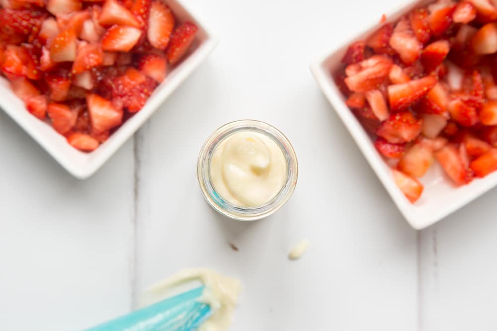 over head view of a small jar with cheesecake mix to the top with two bowls of chopped strawberries at either side and a blue icing bag.