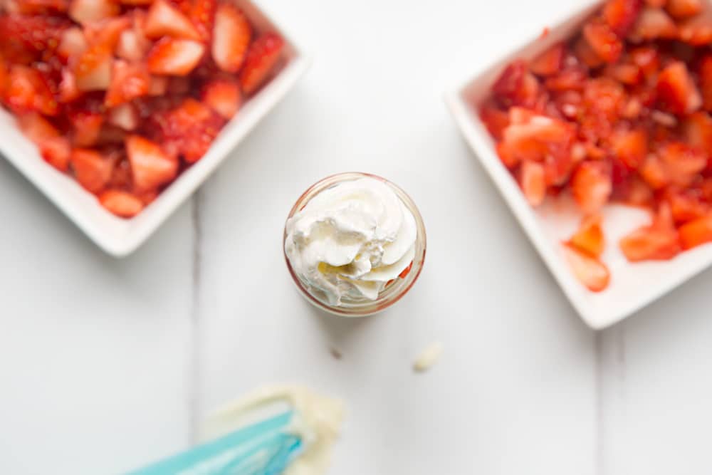 over head view of a small jar with whipped cream topping with two bowls of chopped strawberries at either side and a blue icing bag.