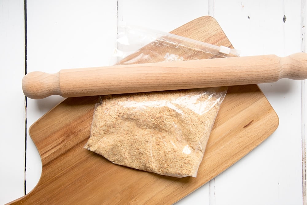 crushed digestive biscuits in a food bag laid on a chopping board with a rolling pin on top.