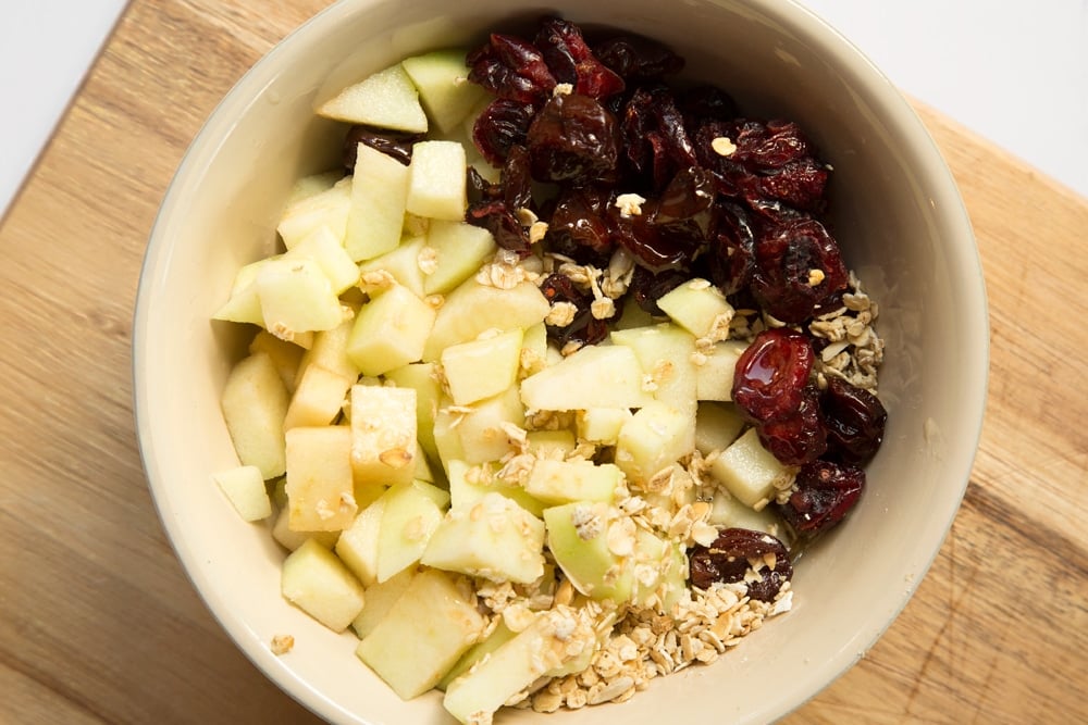 Granola, apples, cherries and honey in a bowl on top of a chopping board.