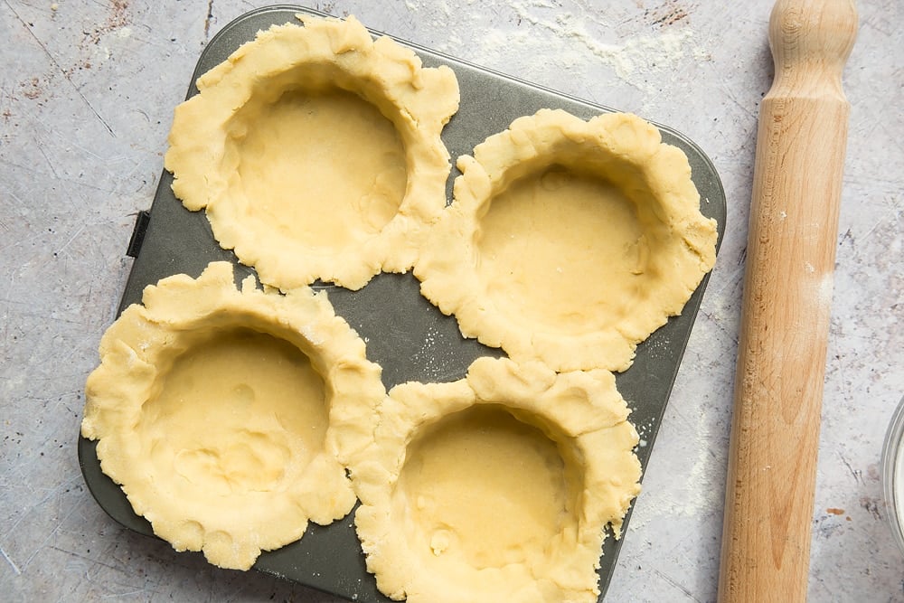 4 raw pastry cases in a metal pastry tray with a rolling pin at the side.