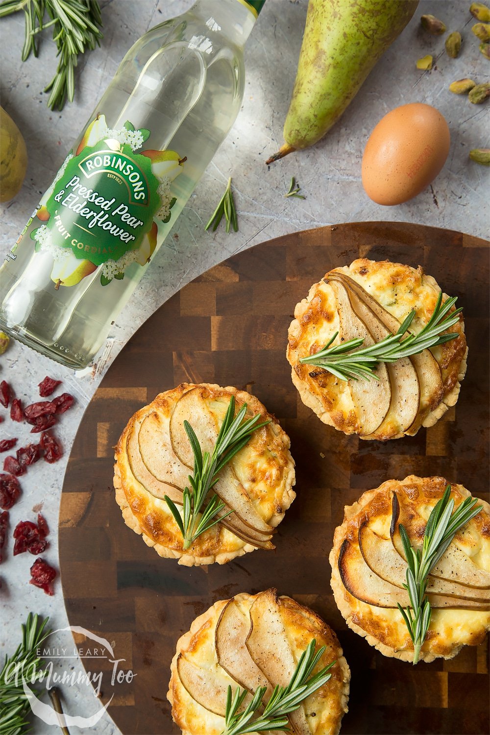 Overhead shot of a set of rosemary goat's cheese tarts with sliced bear with a botte of the Robbinsons pressed pear & elderflower on the side. 