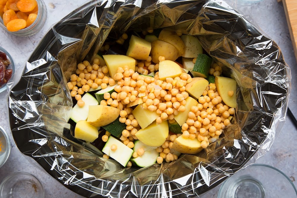 over head shot of a slow cooker with a slow cooker liner and chopped raw vegetable pieces and chickpeas inside.