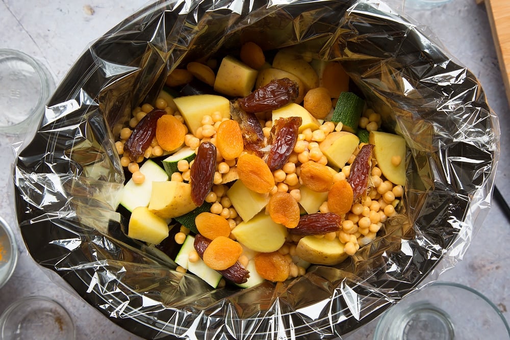 over head shot of a slow cooker with a slow cooker liner and chopped raw vegetable pieces, dates and chickpeas inside.