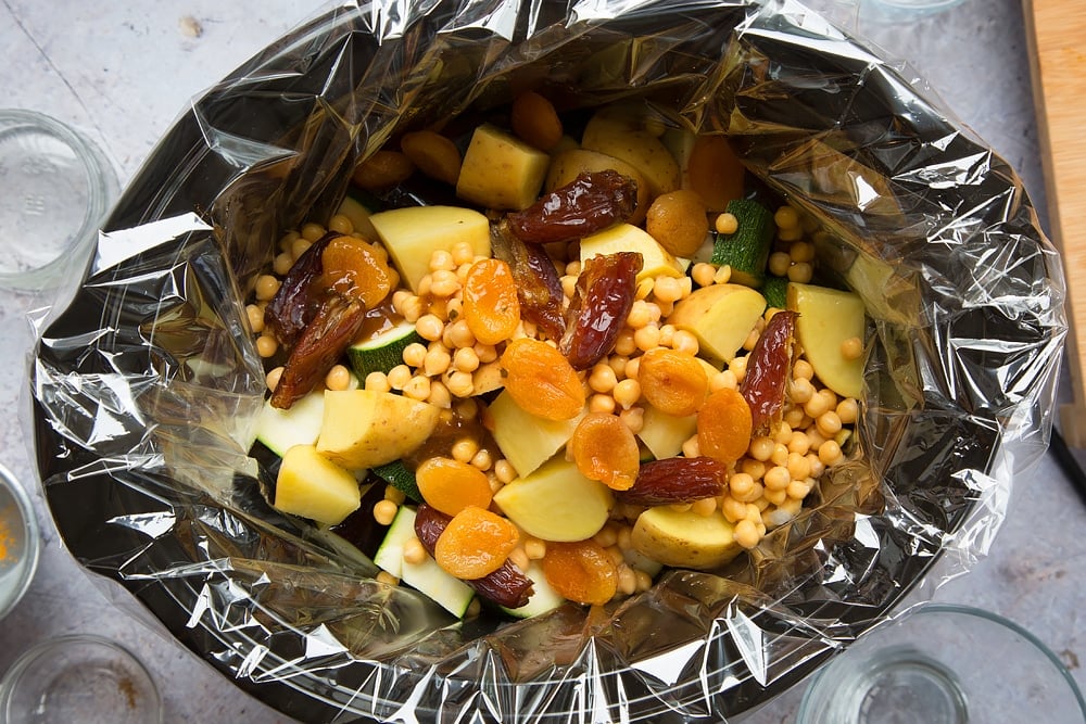 over head shot of a slow cooker with a slow cooker liner and chopped raw vegetable pieces, dates and chickpeas inside.