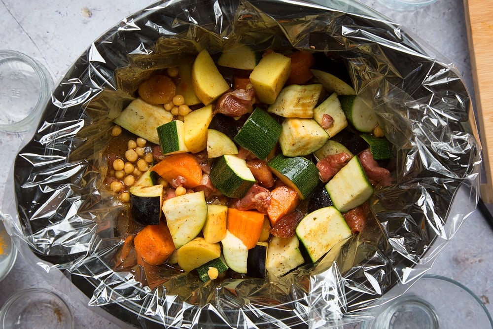 over head shot of a slow cooker with a slow cooker liner and chopped raw vegetable pieces, dates and chickpeas inside with stock.