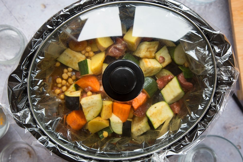 over head shot of a slow cooker with a slow cooker liner and chopped raw vegetable pieces, dates and chickpeas inside with lid on top.