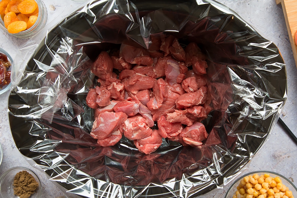 over head shot of a slow cooker with a slow cooker liner and chopped raw lamb pieces inside.