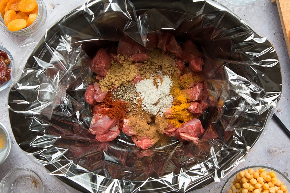 over head shot of a slow cooker with a slow cooker liner and chopped raw lamb pieces inside with spices.