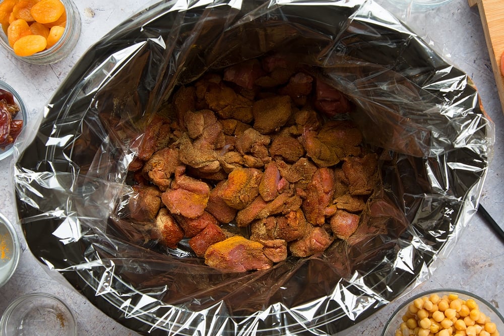 over head shot of a slow cooker with a slow cooker liner and chopped raw lamb pieces inside smothered with spices.