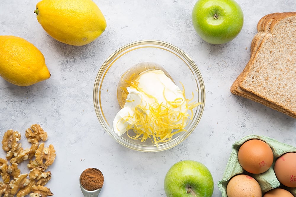 mascarpone, lemon zest, and honey in a bowl surrounded by apples, lemons, eggs and bread.