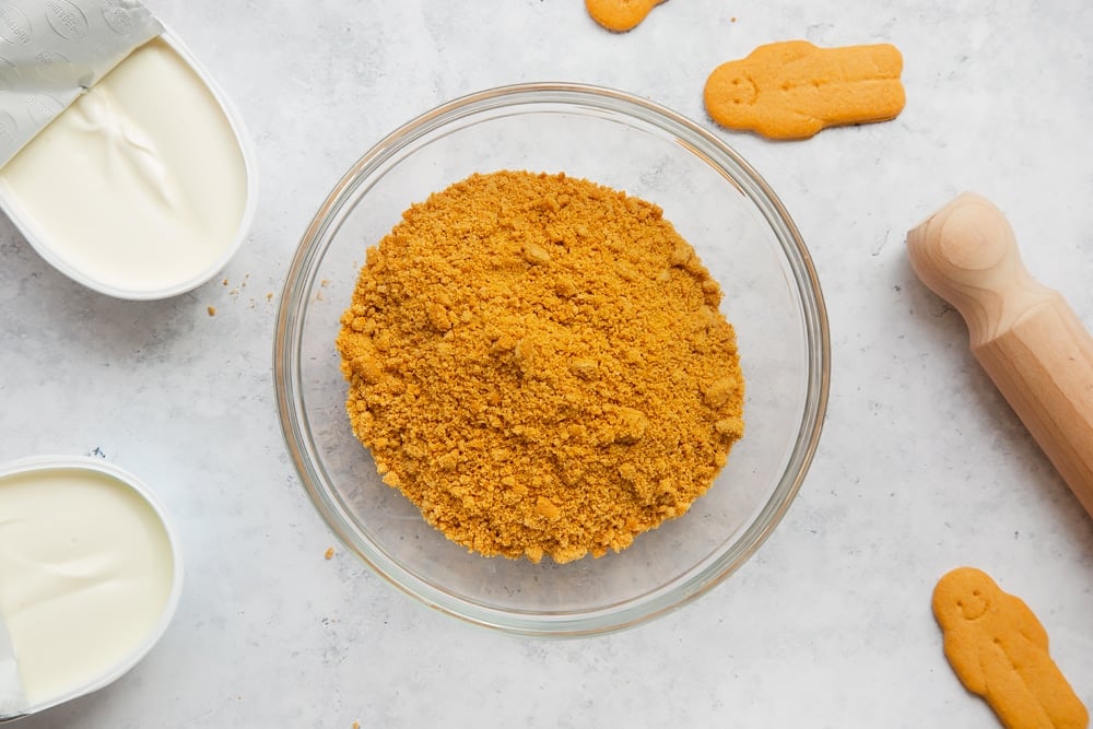 Overhead shot of crushed biscuits in a clear bowl with a wooden rolling pin on the side