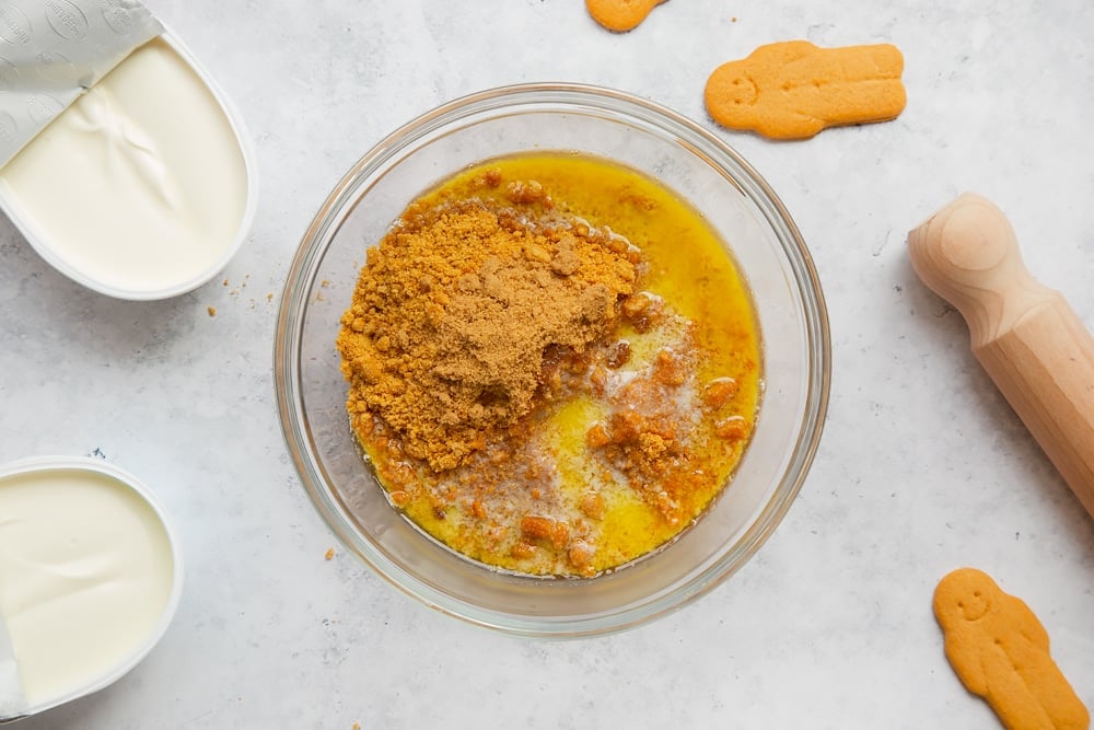 Overhead shot of crushed biscuits, sugar and melted butter in a clear bowl with a cream cheese on the side