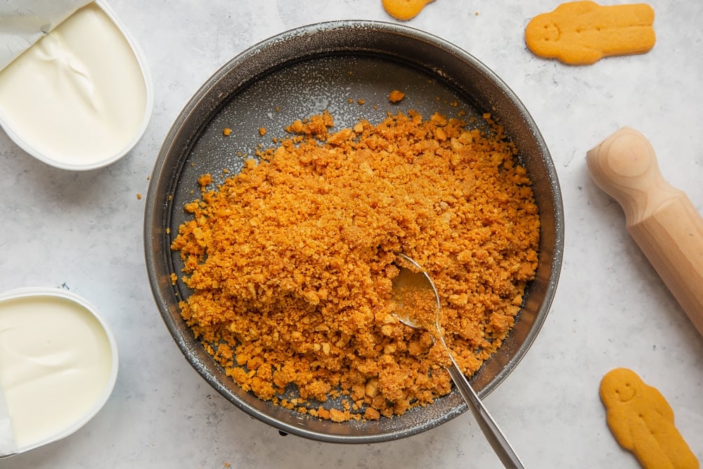 Overhead shot of crushed biscuits mix in a large springform cake tin with a spoon