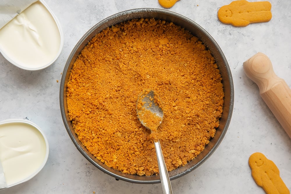 Overhead shot of crushed biscuits mix in a large springform cake tin with a spoon