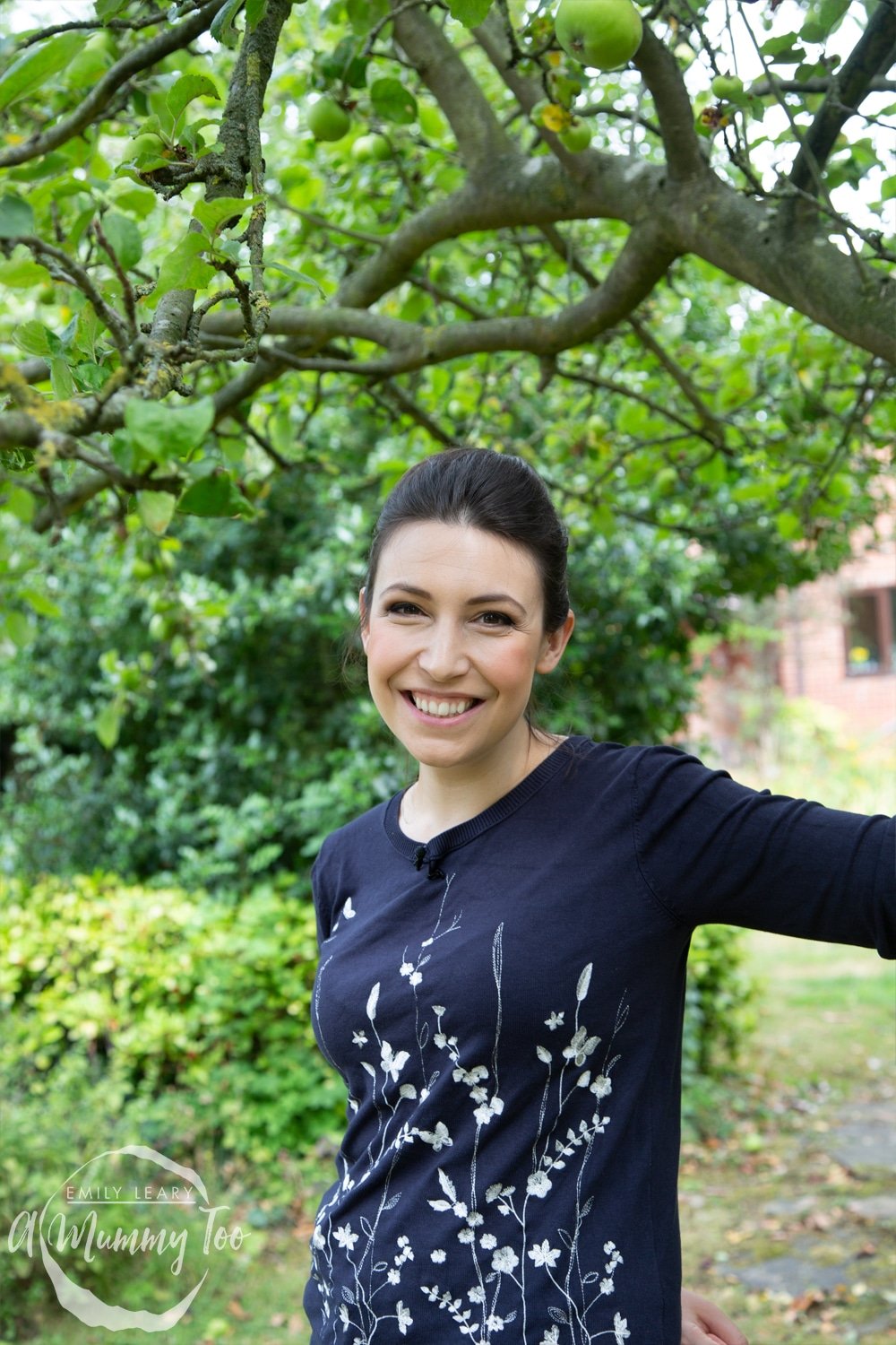 a young woman with dark hair tied up wearing a navy blue floral long sleeve top stood outside in a garden. Emily Leary A Mummy Too