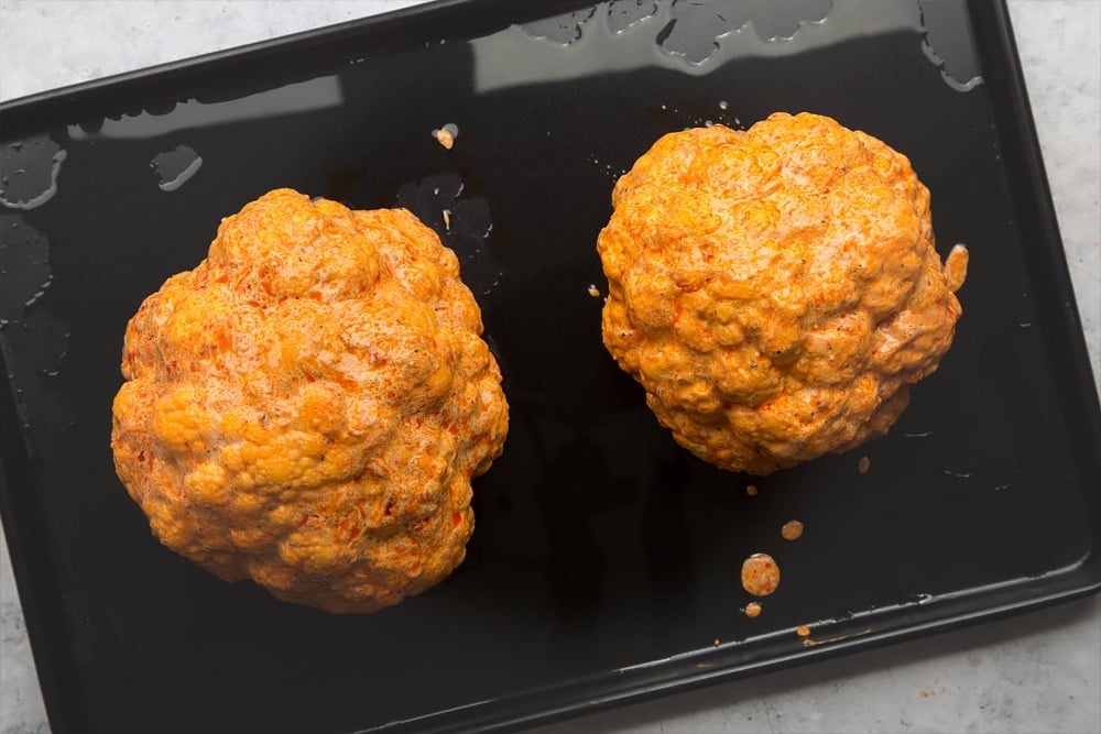 Two cauliflowers covered in harissa sauce on a baking tray