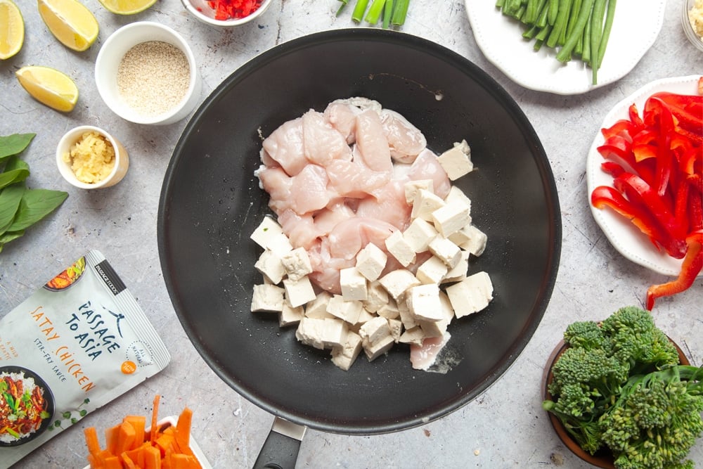 Diced chicken and tofu in a black wok surrounded by vegetables on white plates and passage to asia sauce packet.