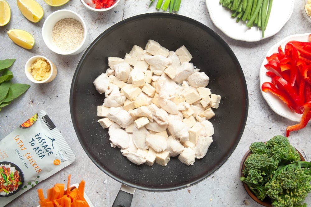 Chicken and tofu in a stir fry pan surrounded by vegetables and ingredients.