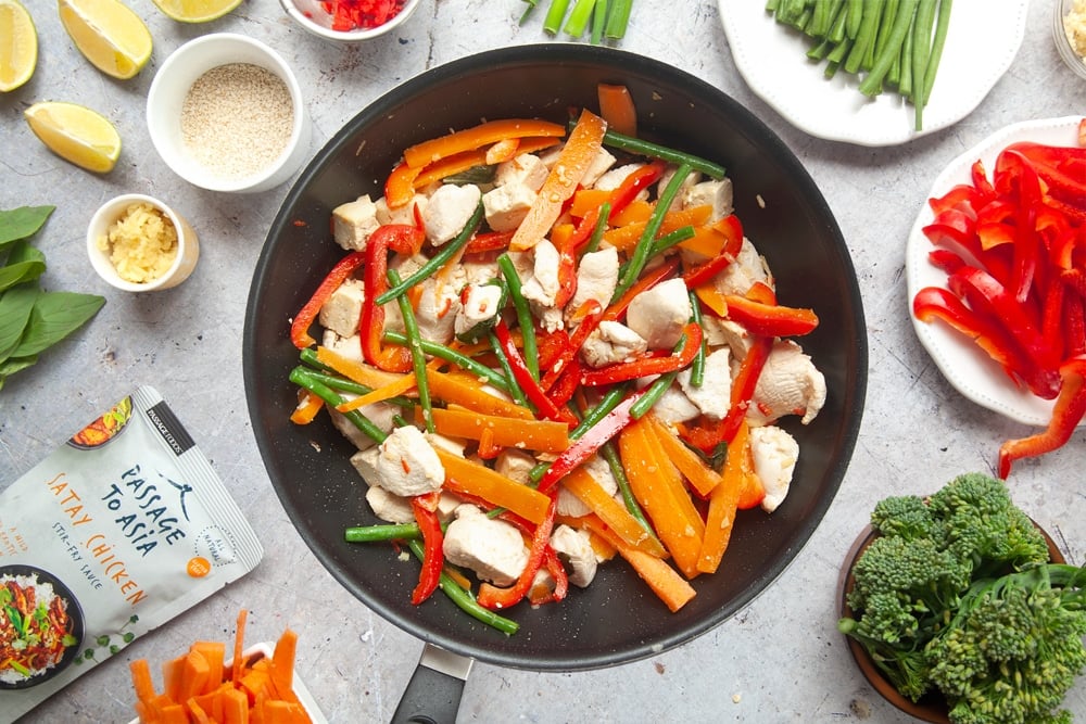 Cooking the chicken and tofu satay stir-fry in a black wok surrounded by cut vegetables and ingredients.