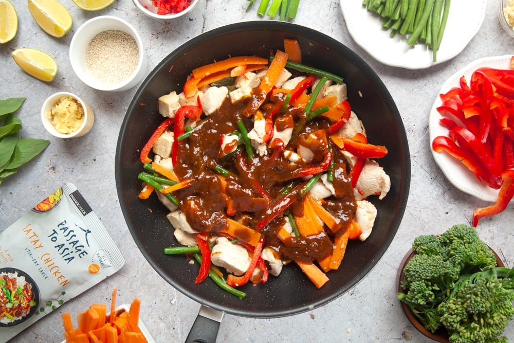 Chicken and tofu stir fry topped with satay chicken sauce surrounded by cut vegetables and ingredients.