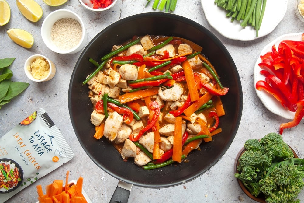 Cooking chicken and tofu satay stir-fry in a wok surrounded by cut vegetables and ingredients.