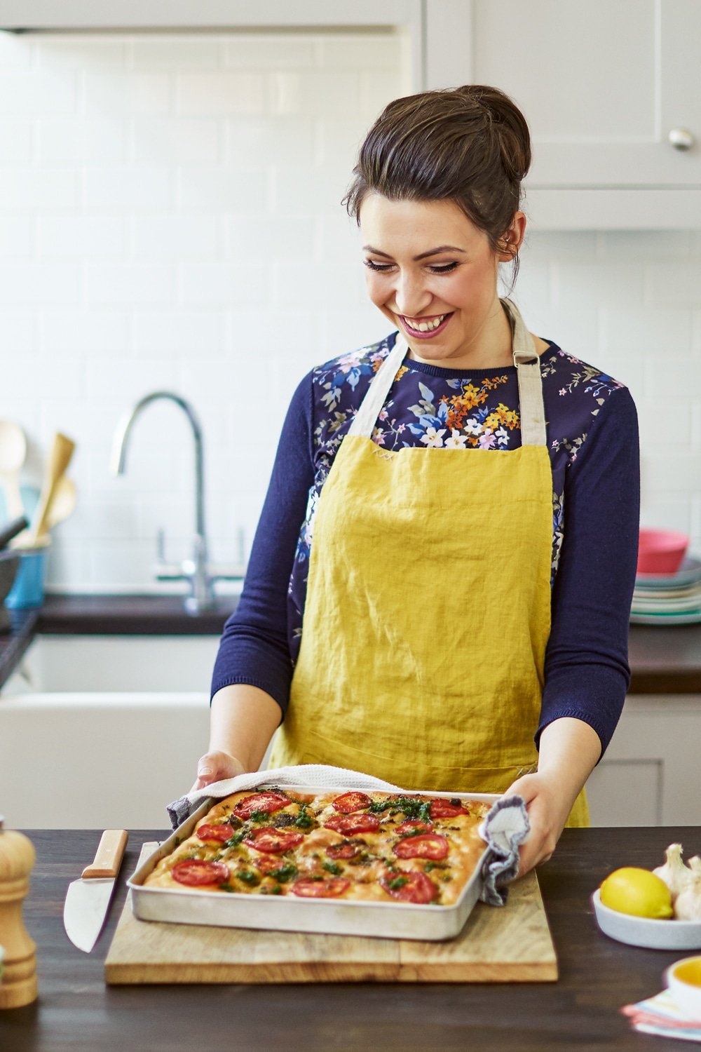 Emily Leary in a blue long sleeved t-shirt with a yellow apron holding one of her famous recipes inside a sheet pan on top of a wooden chopping board in a kitchen.