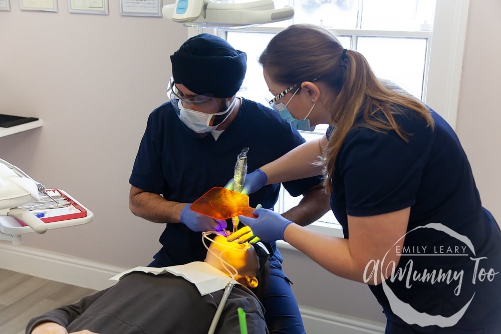 view of a dentist studio with a young woman laid out on a dentist chair with two dentists.