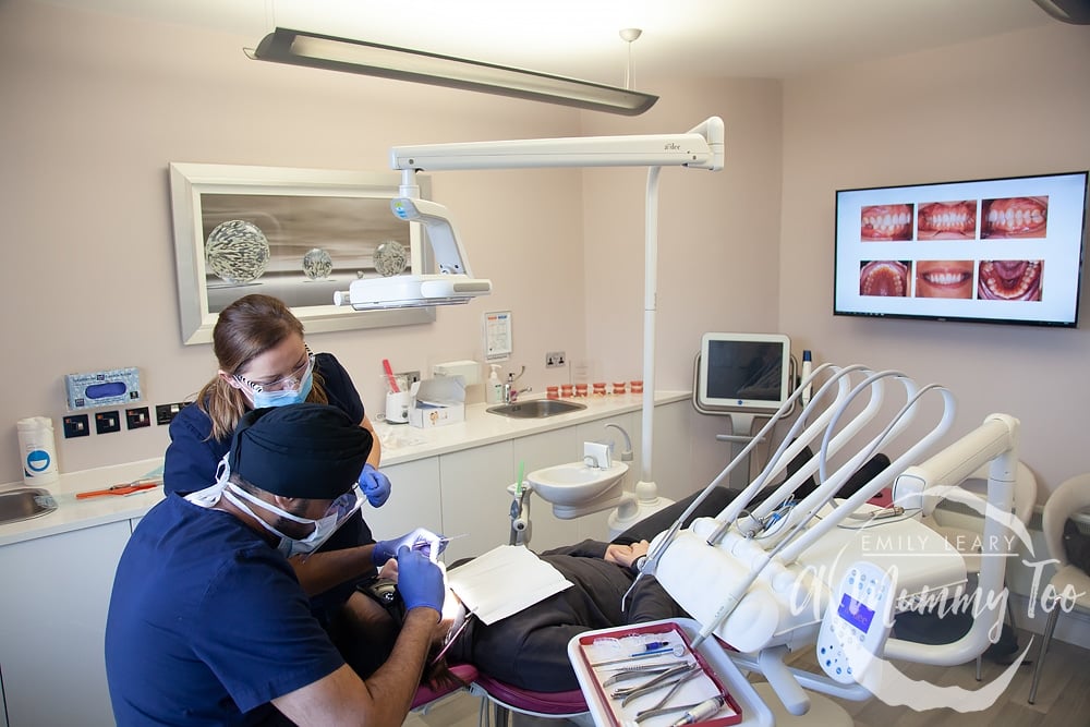 view of a dentist studio with a young woman laid out on a dentist chair with two dentists.