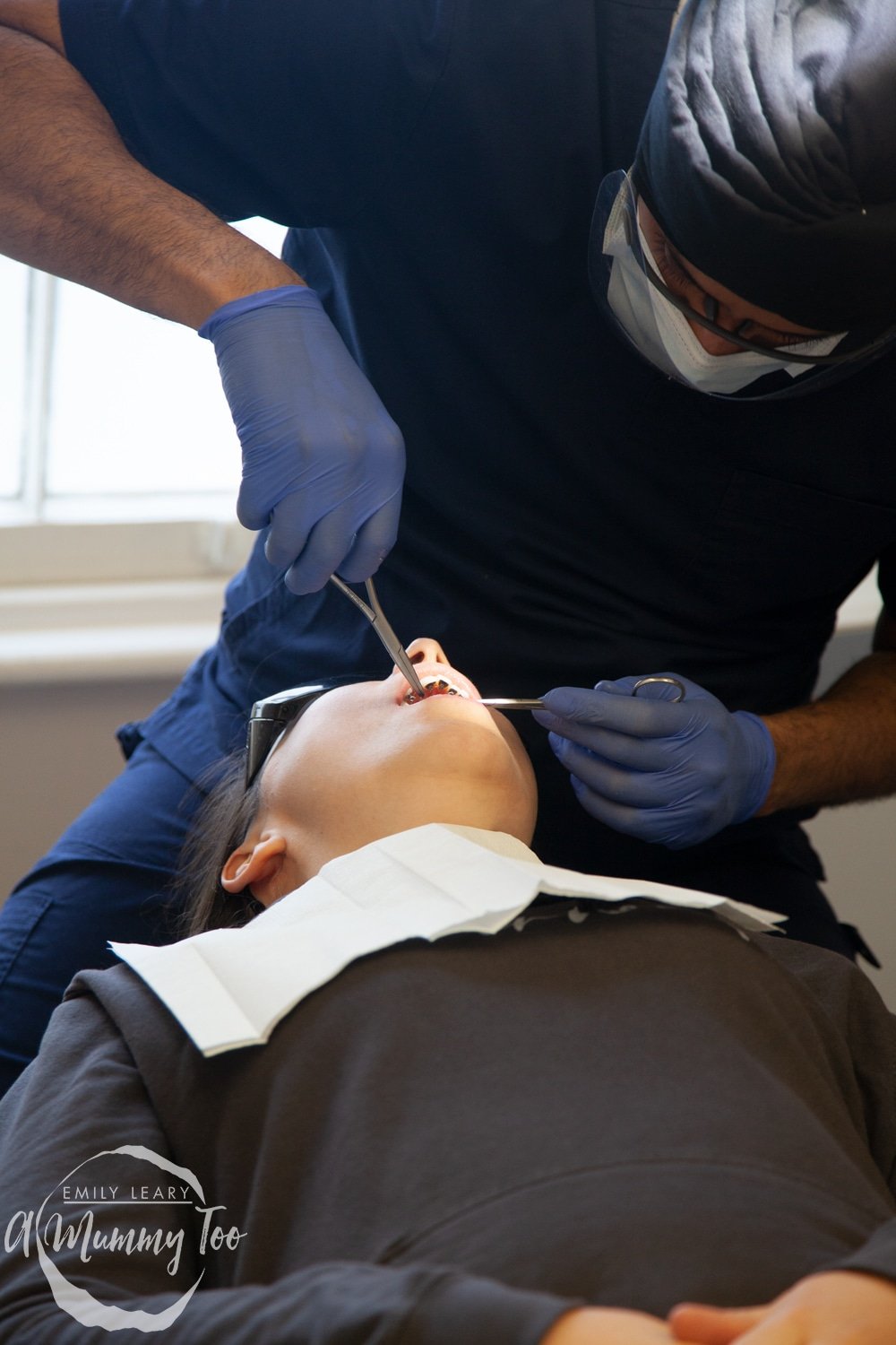 a young woman laid on a dentist chair while a dentist works on her teeth.