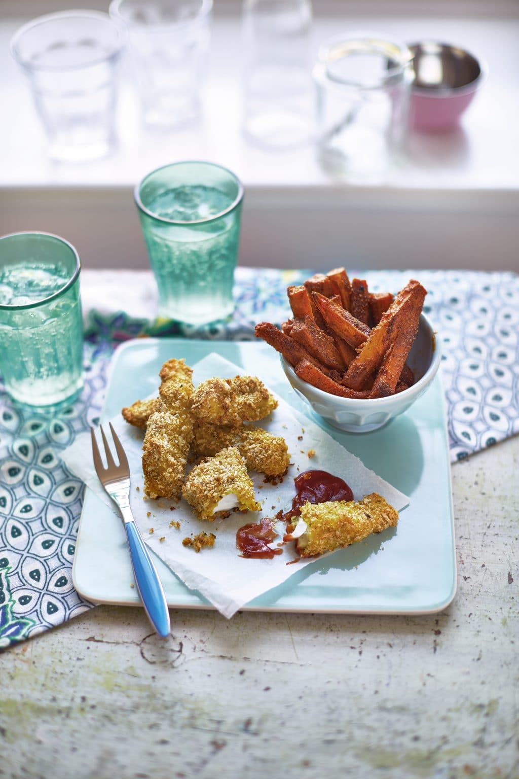 Front angle shot of curried fish fingers with ketchup on the side and sweet potato fries served on a teal plate