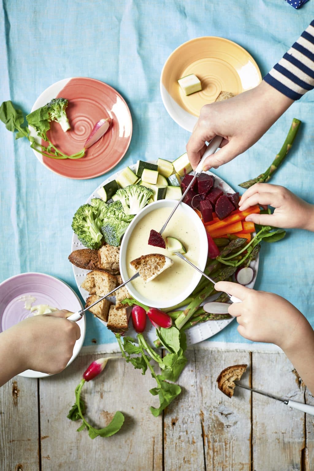 Overhead shot of hands dipping bread and veggies with fondue forks in white sauce from Healthier Fondue with Veggies served in a white plate