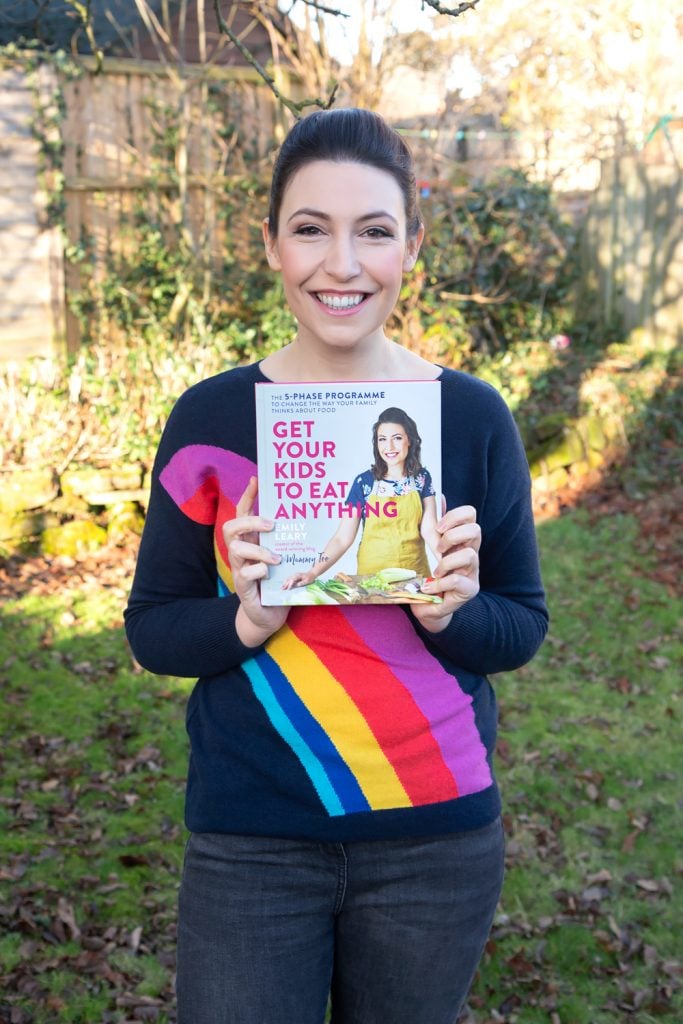 Front angle shot of Emily Leary smiling holding up a cookbook Get Your Kids to Eat Anything