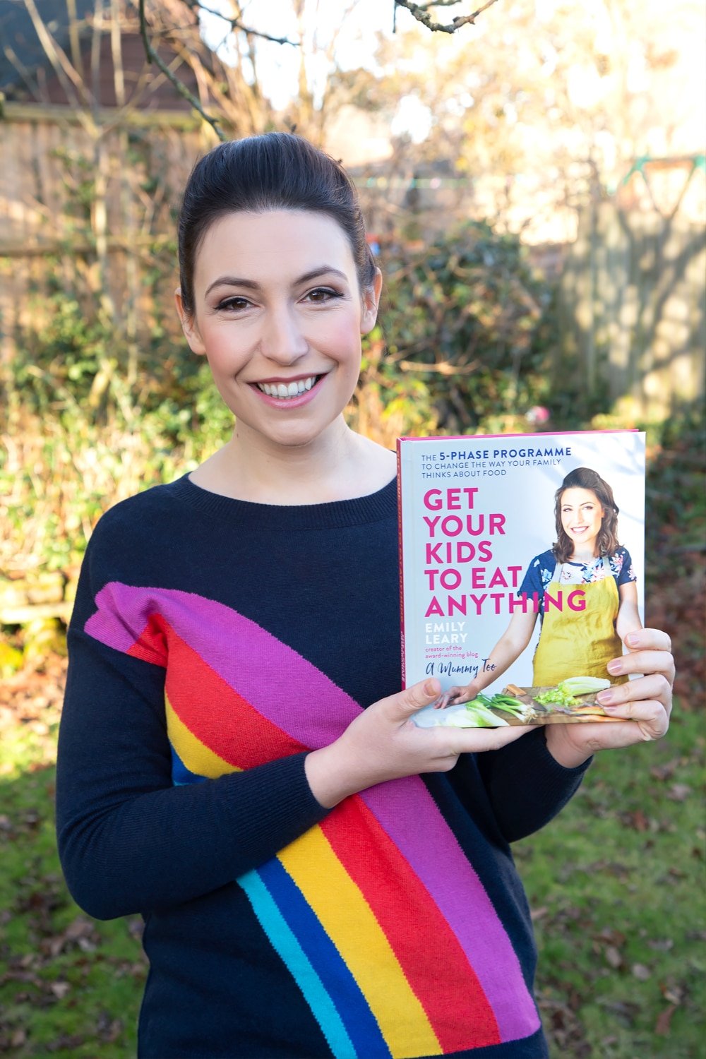 Front angle shot of Emily Leary smiling holding up a cookbook Get Your Kids to Eat Anything