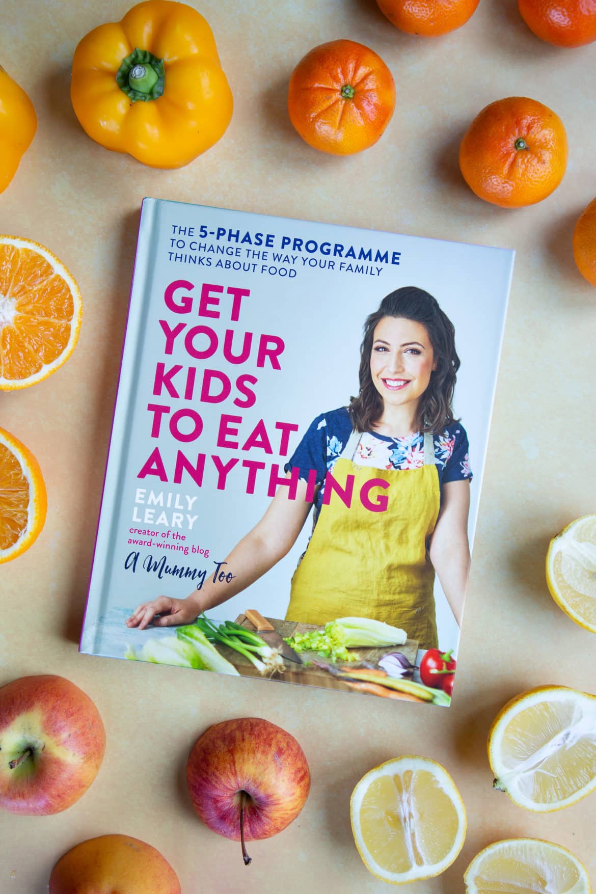 Photograph of the hardcover book: Get Your Kids To Eat Anything by Emily Leary. The book is on a pale yellow background and is surrounded by apples, halved lemons, tangerines, a yellow pepper and halved oranges. 