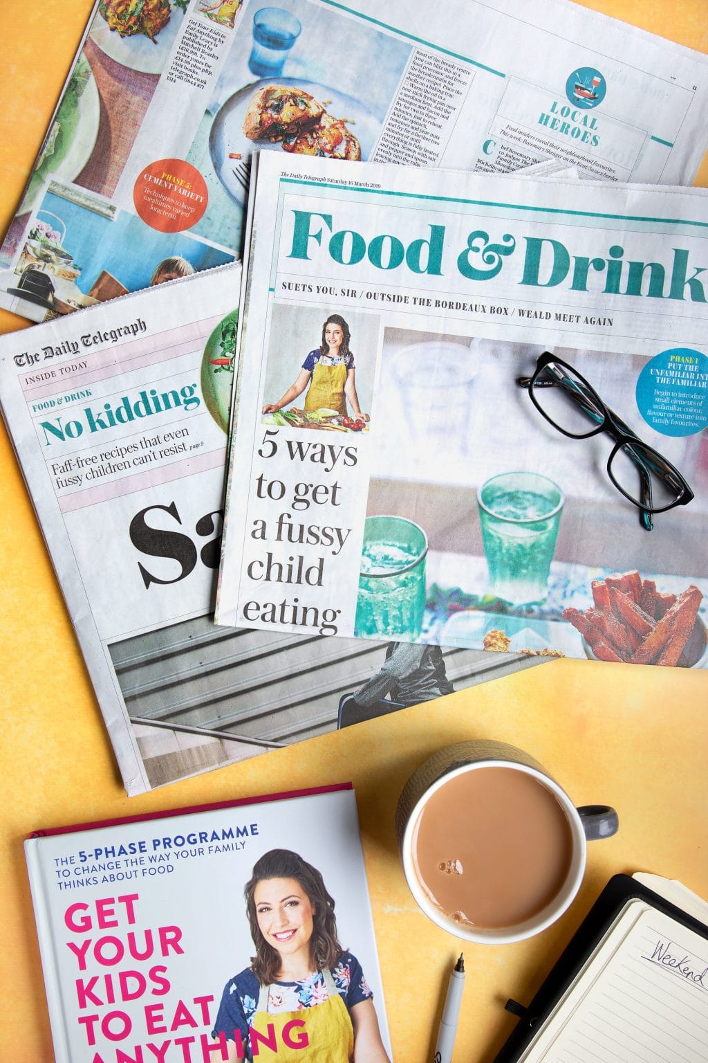 Overhead shot of glasses, newspapers, coffee, and Get Your Kids to Eat Anything cookbook